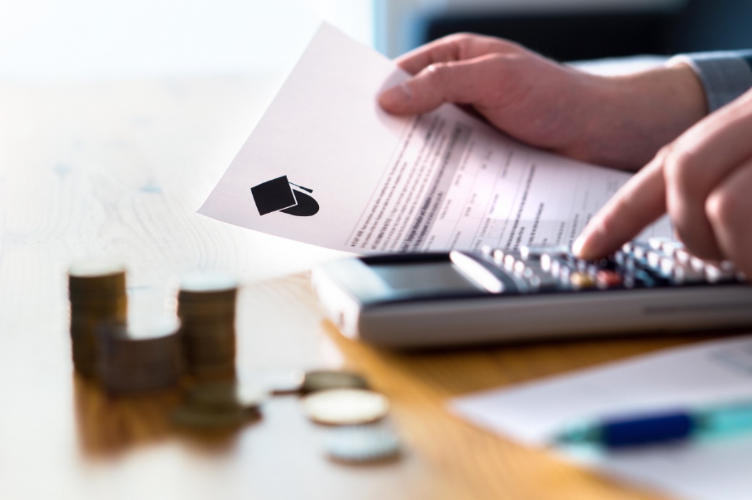 Individual using a calculator to manage finances, holding a document with a graduation cap icon, coins stacked on the table.