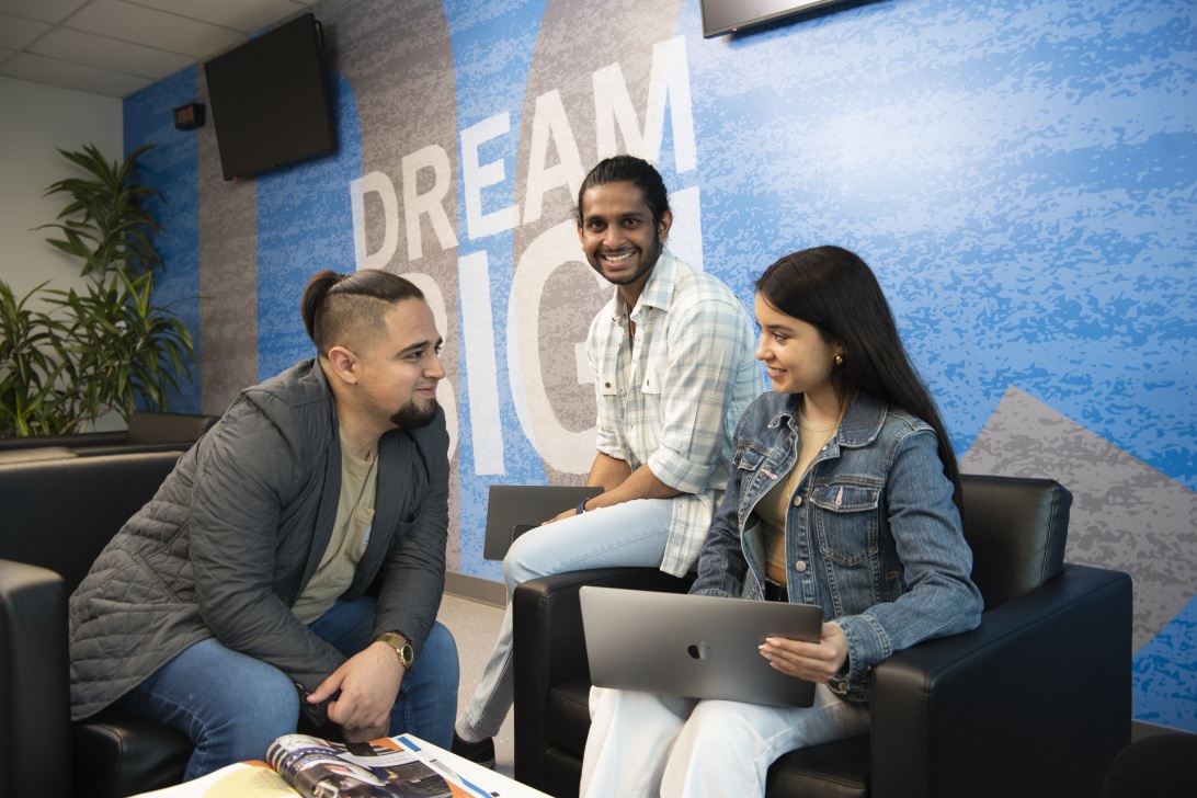 Three people sit in a lounge area talking; one uses a laptop and a magazine is on the table. A "DREAM BIG" mural is behind them.