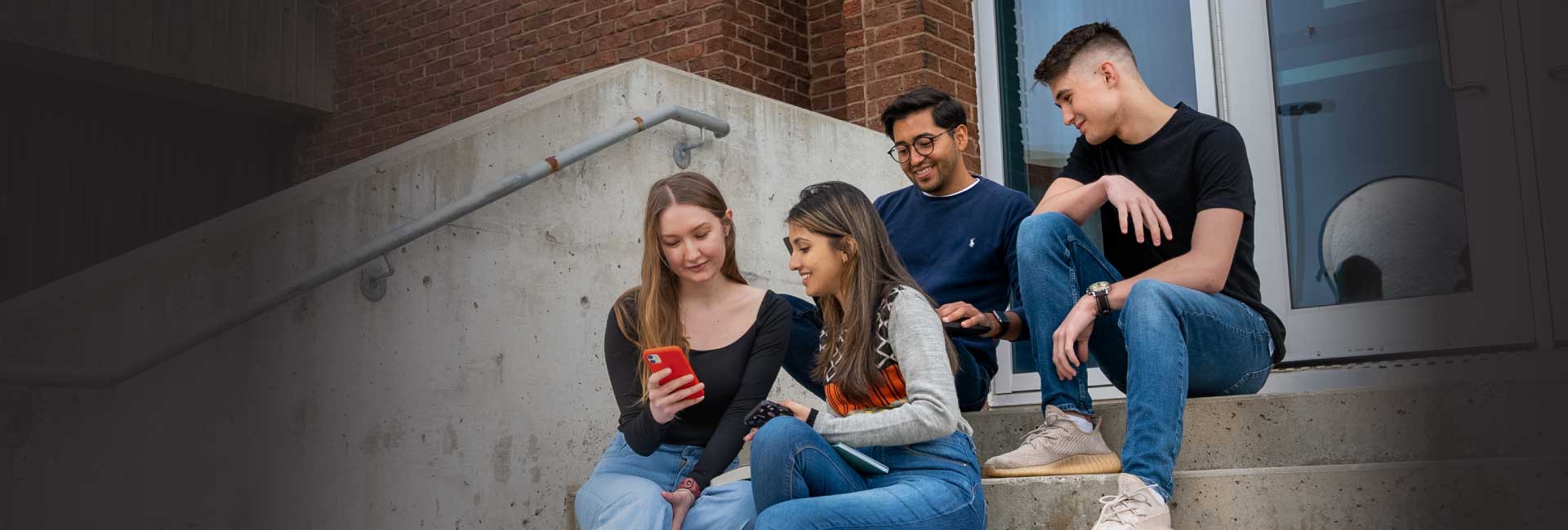A group of students sit on stairs at the entrance of a building, all looking at a smart phone held by one of the students.