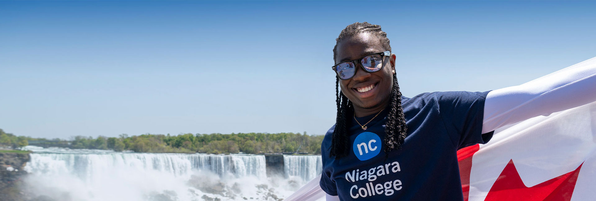 A student wearing a Niagara College t-shirt, posing for a photo at Niagara Falls while holding a Canadian flag.